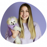 A picture of our groomer, Gabby. She is holding a white fluffy dog in a purple coat.